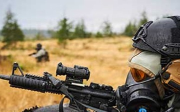 Military personnel wearing protective respiratory equipment and carrying rifles participate in a training exercise.