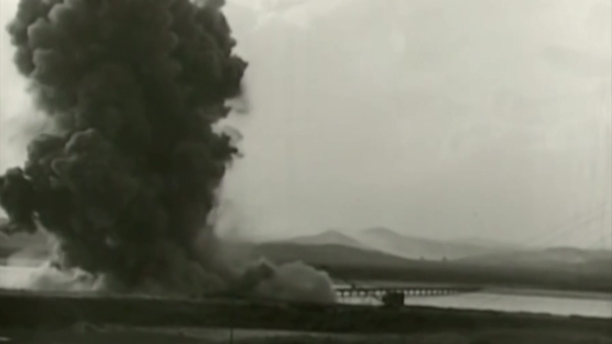 Black smoke billows up near a body of water, as if from an explosion. 