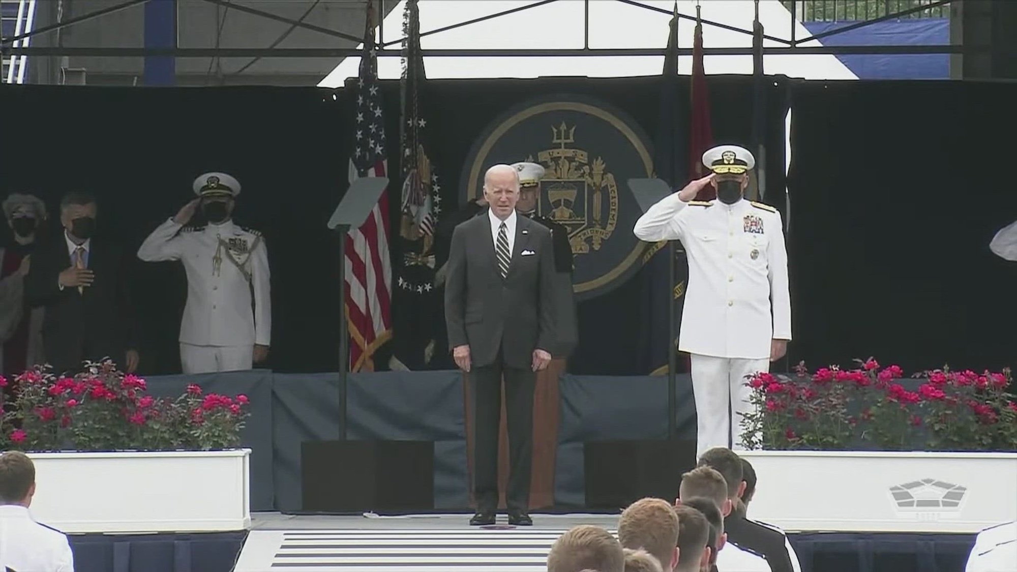 President Joe Biden stands on outdoor stage with saluting sailors. 
