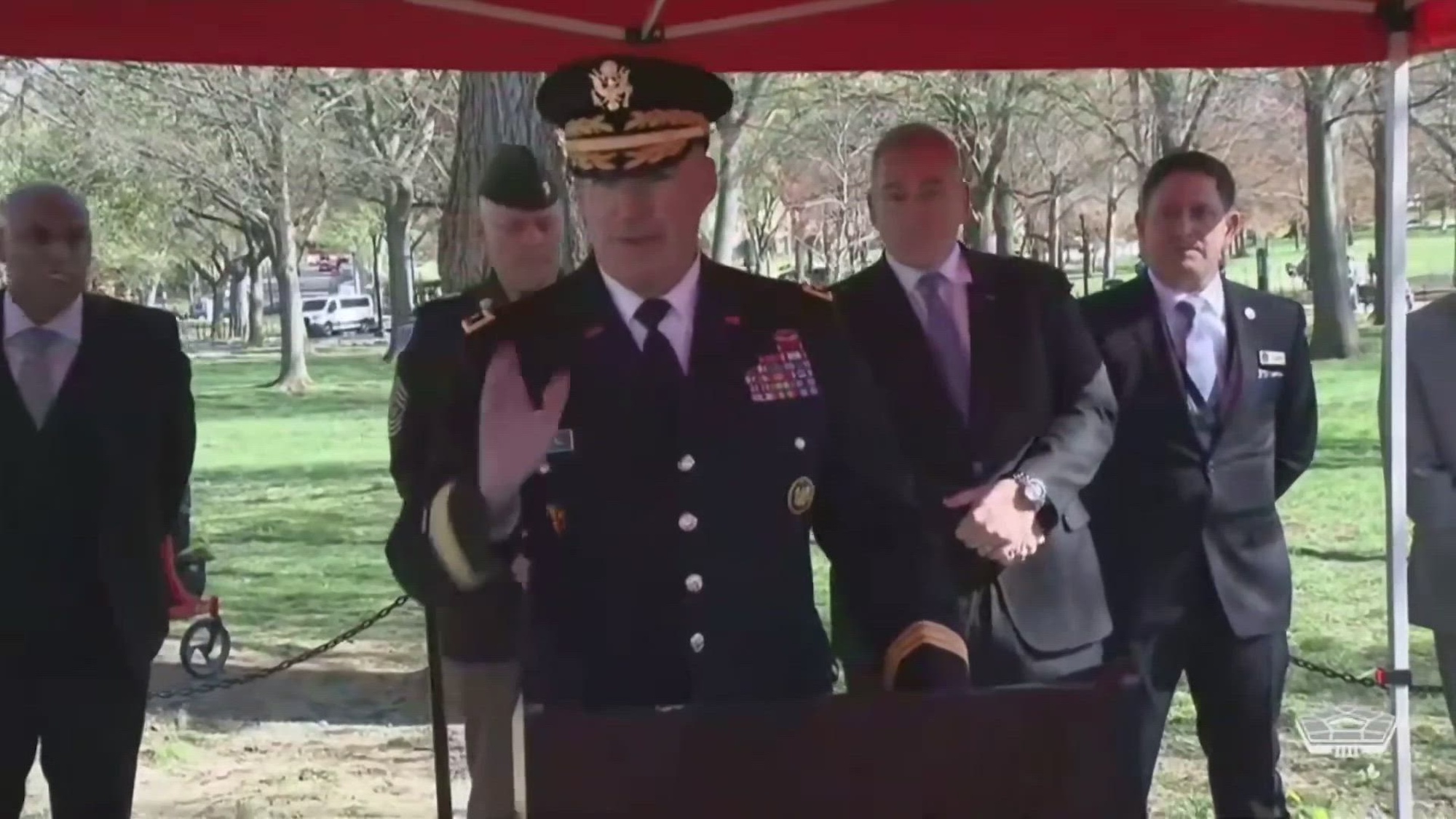 A service member speaks at a lectern outdoors as other officials stand nearby. 
