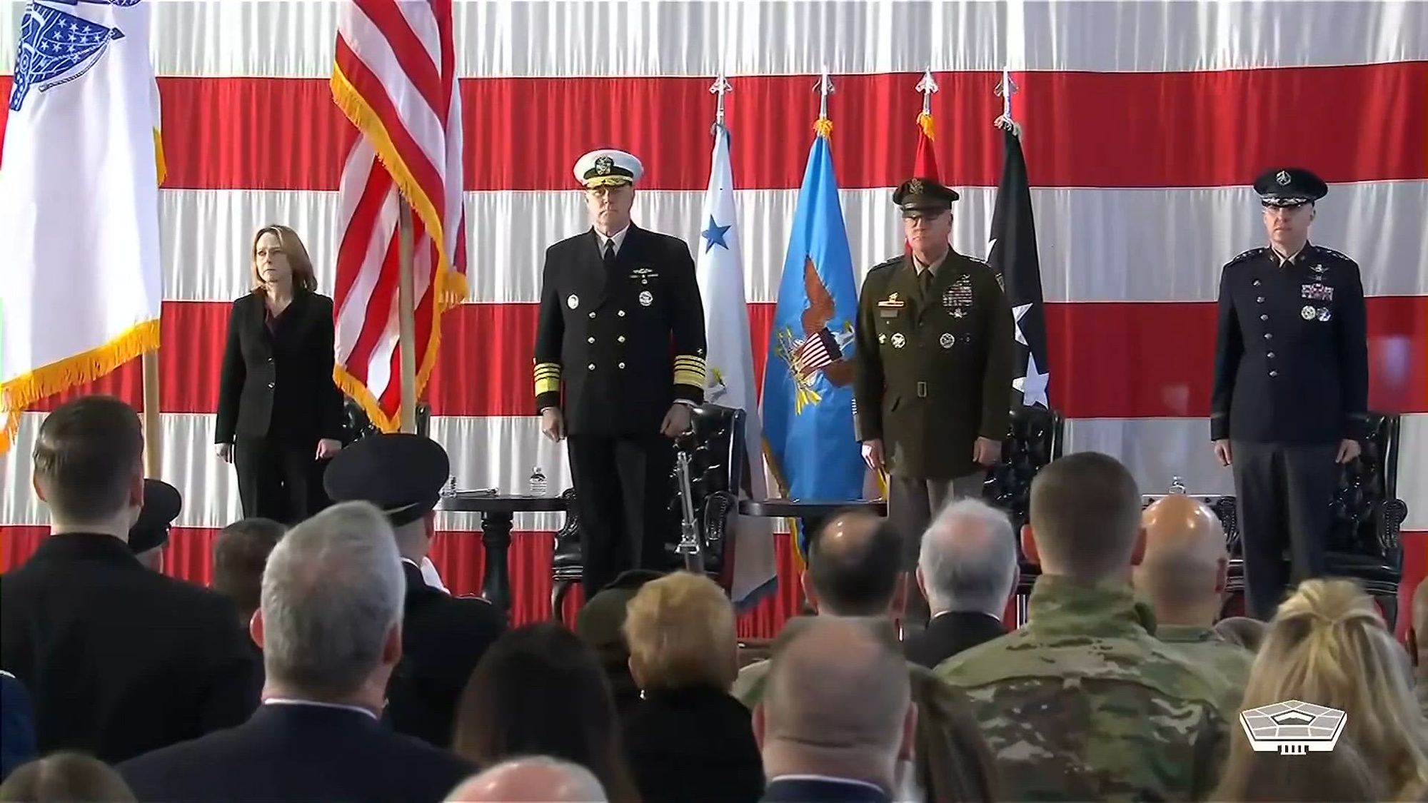 Service members and Deputy Secretary Hicks stand on a flag-draped stage facing an audience.