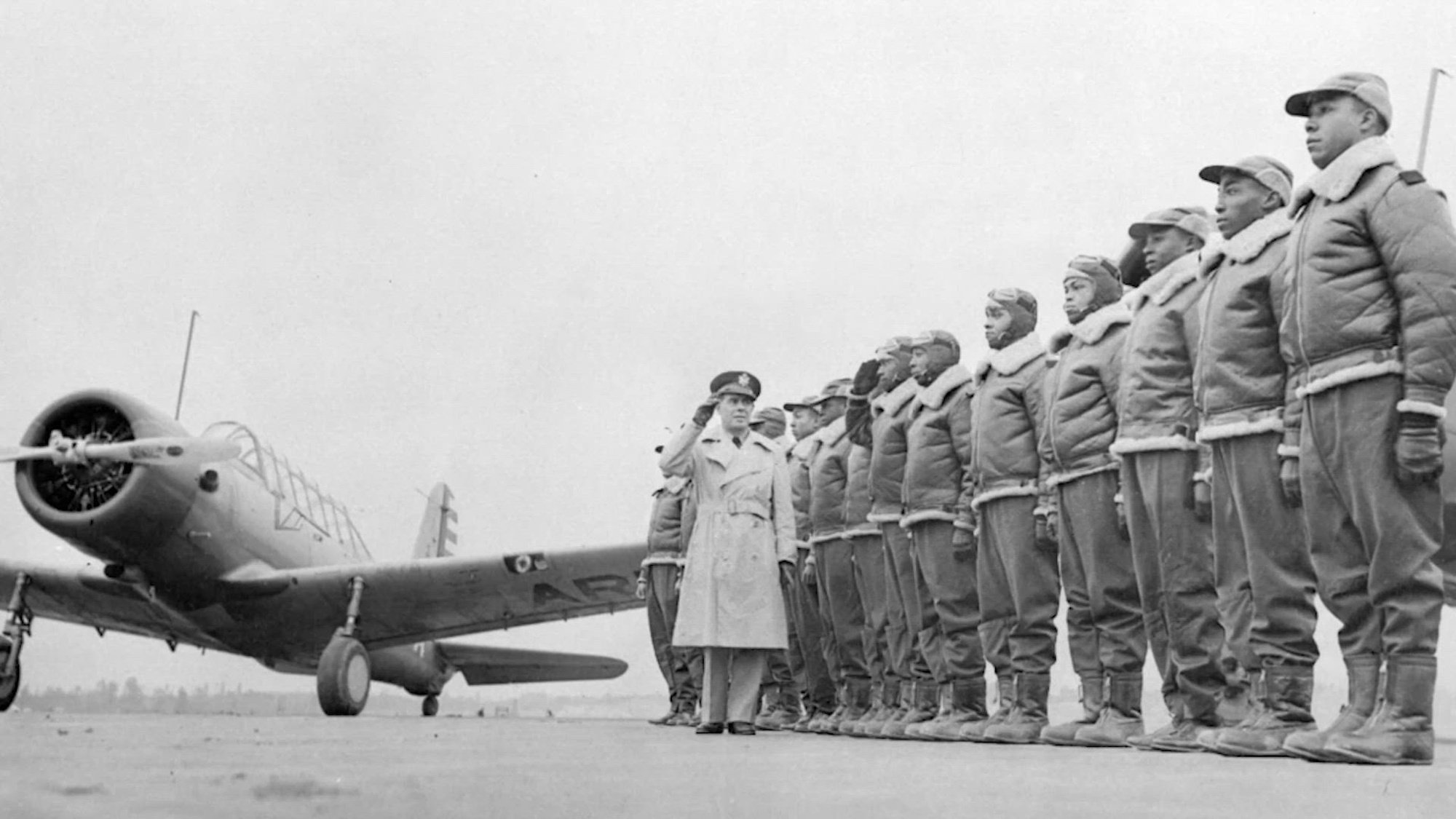 Vintage black and white video of African American Airmen standing in rank at attention adjacent to a P-51 Mustang aircraft.