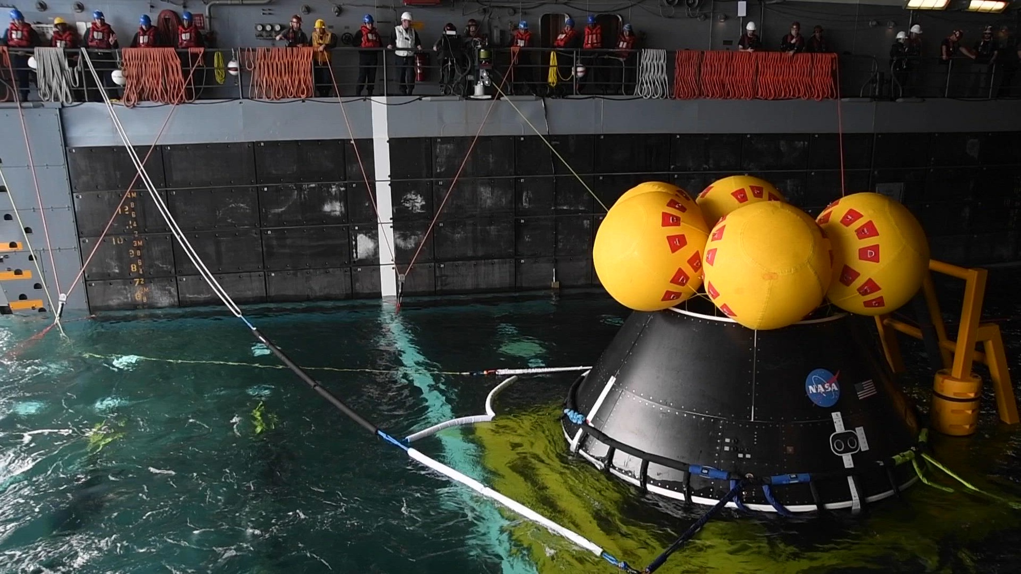 View of a space capsule with yellow spheres attached to in a ship's well deck. 