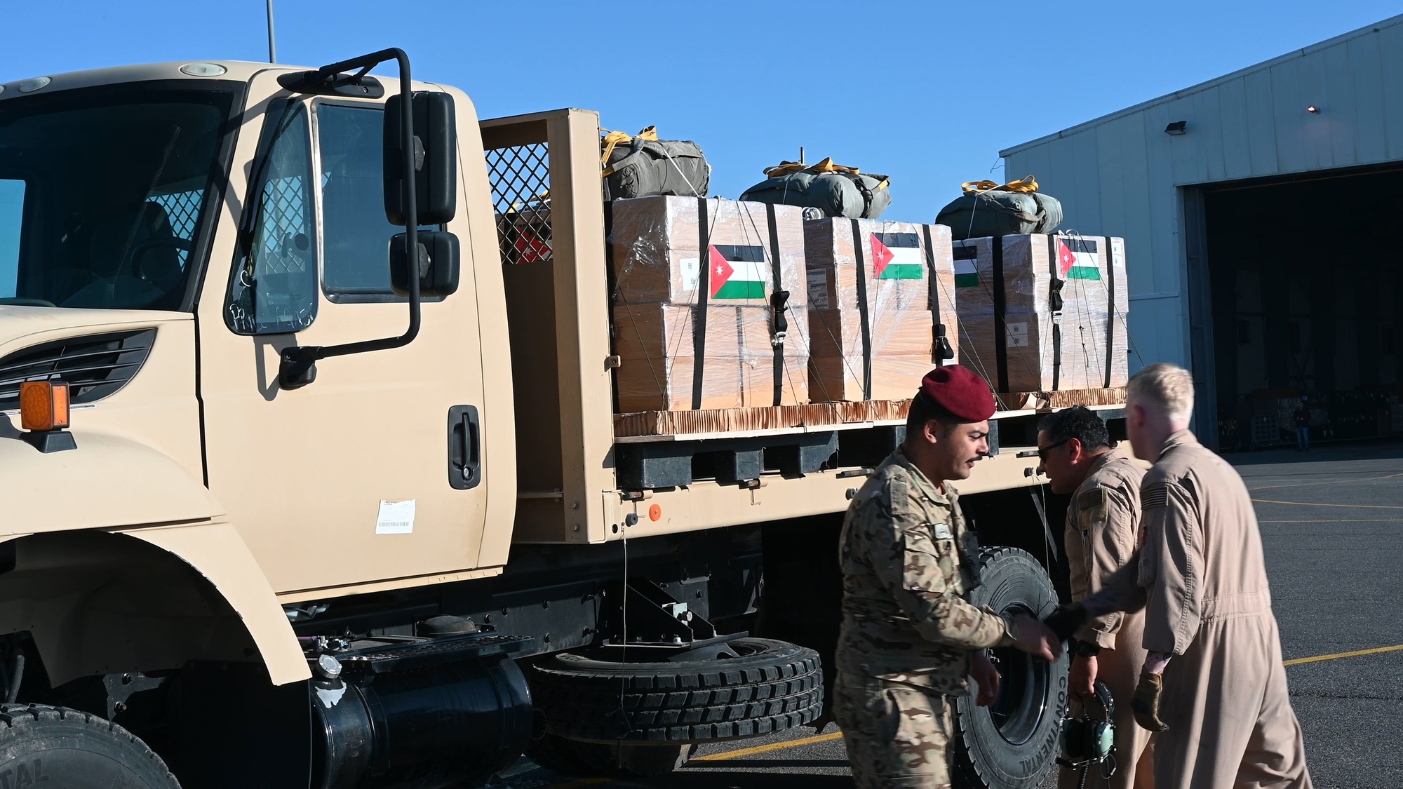 Service members stand next to a truck with pallets holding boxes with the Palestinian flag taped on with a hangar in the background.