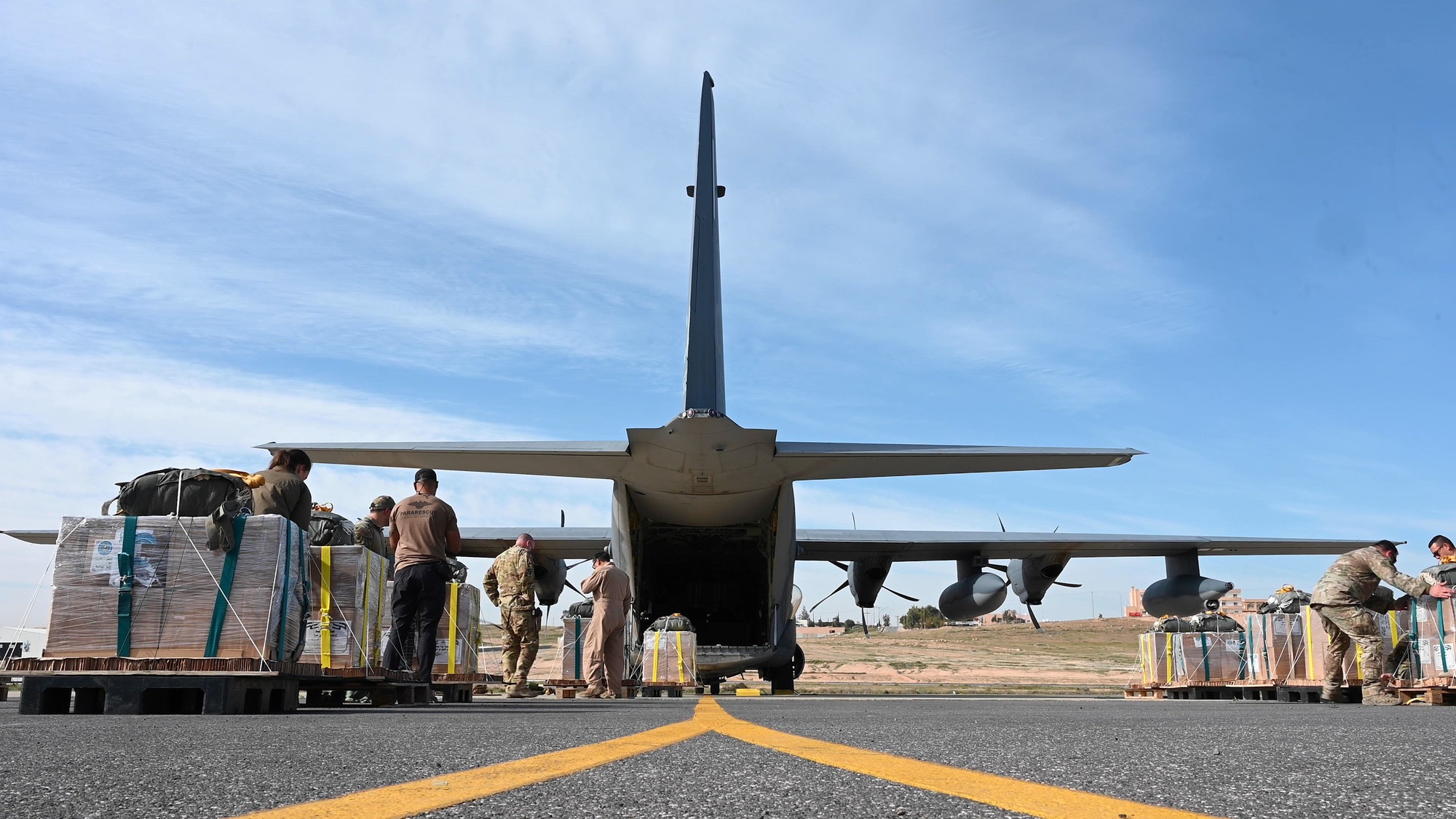 Pallets of aid sit on a runway with airmen nearby while an aircraft's back is open to load.
