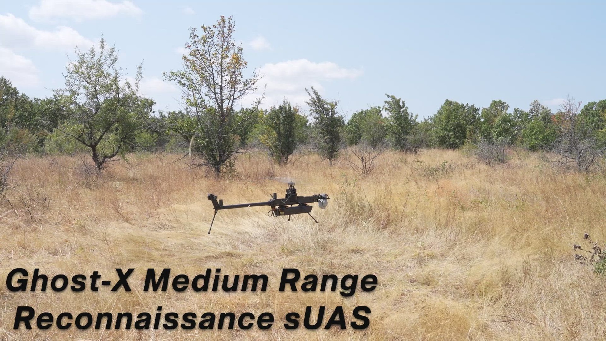 A drone flies over a brown grassy field with green trees and sky in the background