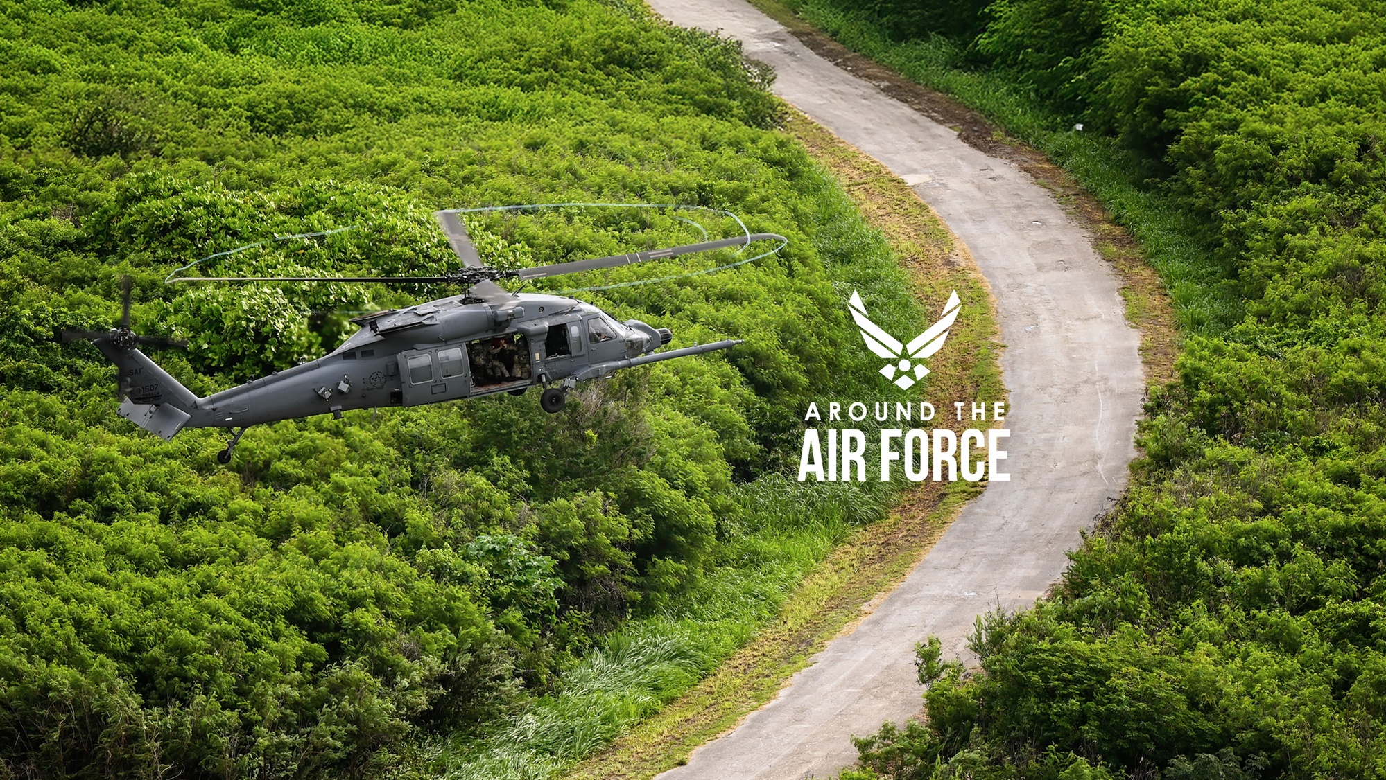 A military helicopter is hovering above a field with a paved road. 