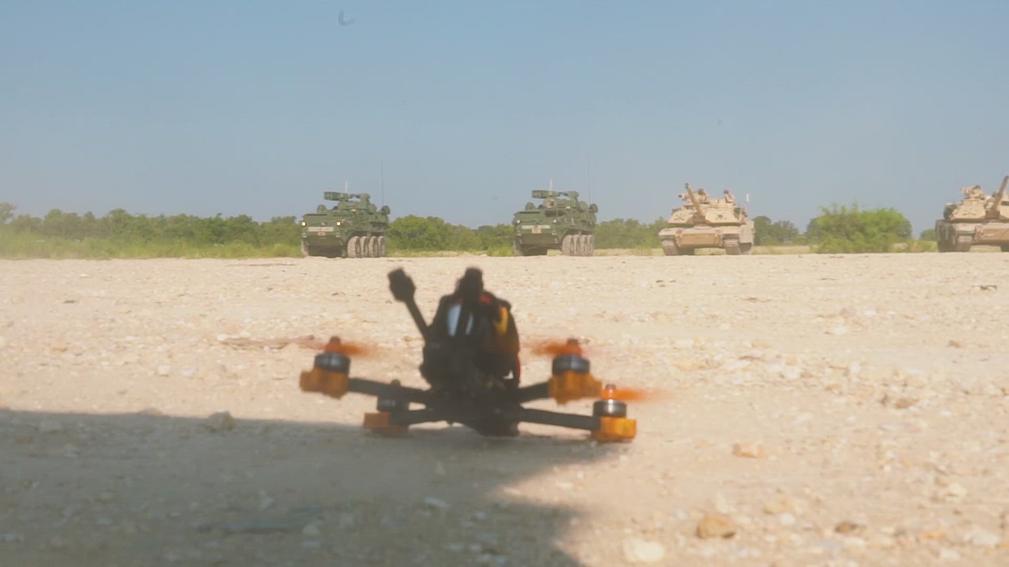 A drone sits on a dirt field; military vehicles are in the background.