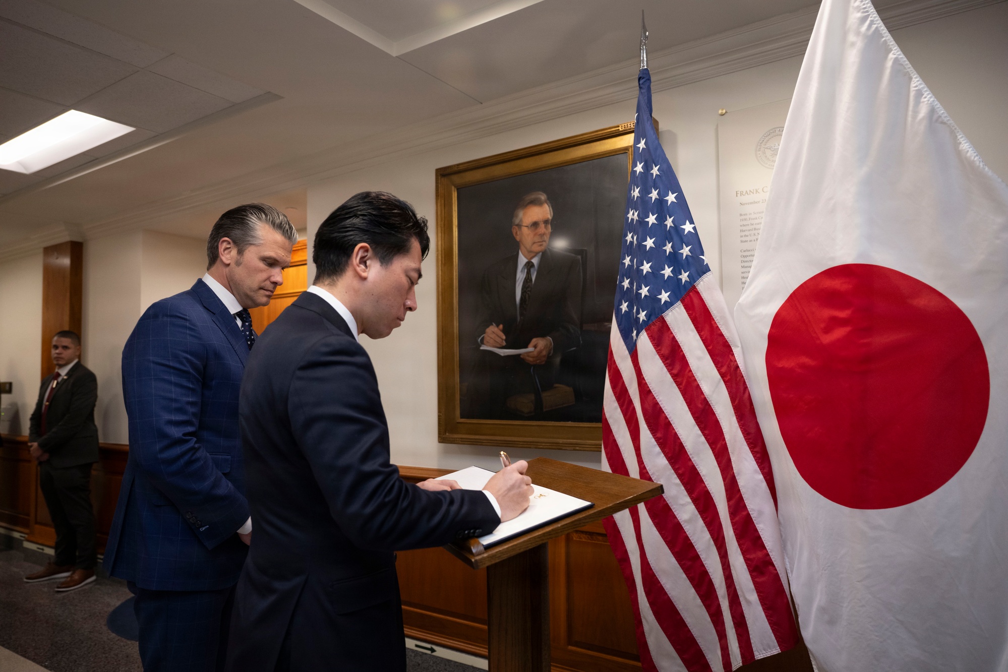 Japanese Minister of Defense Shinjirō Koizumi signs the Pentagon guest book next to Secretary of War Pete Hegseth and the American and Japanese flags.