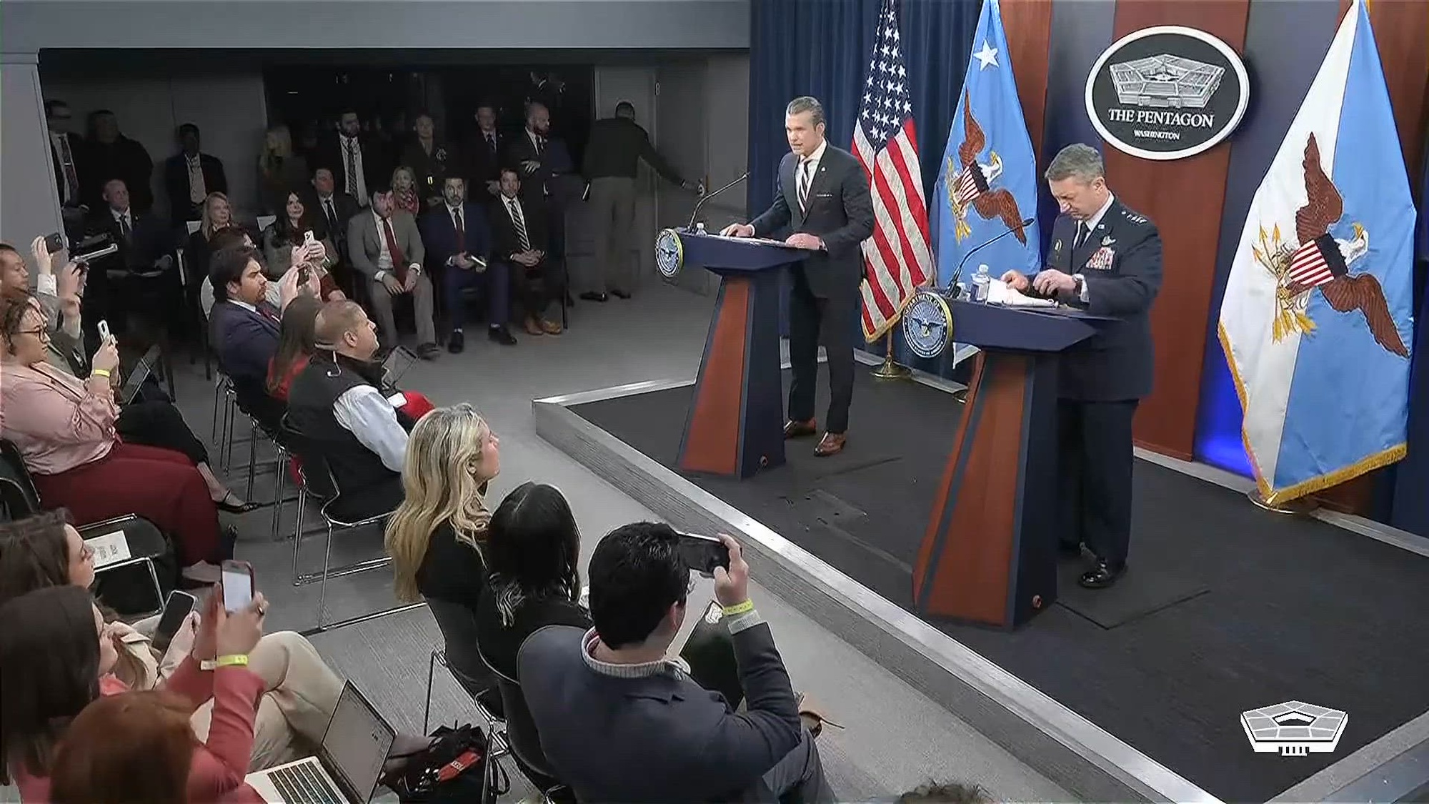 Secretary of War Pete Hegseth and Air Force Gen. Dan Caine stand at two lecterns facing a seated audience.