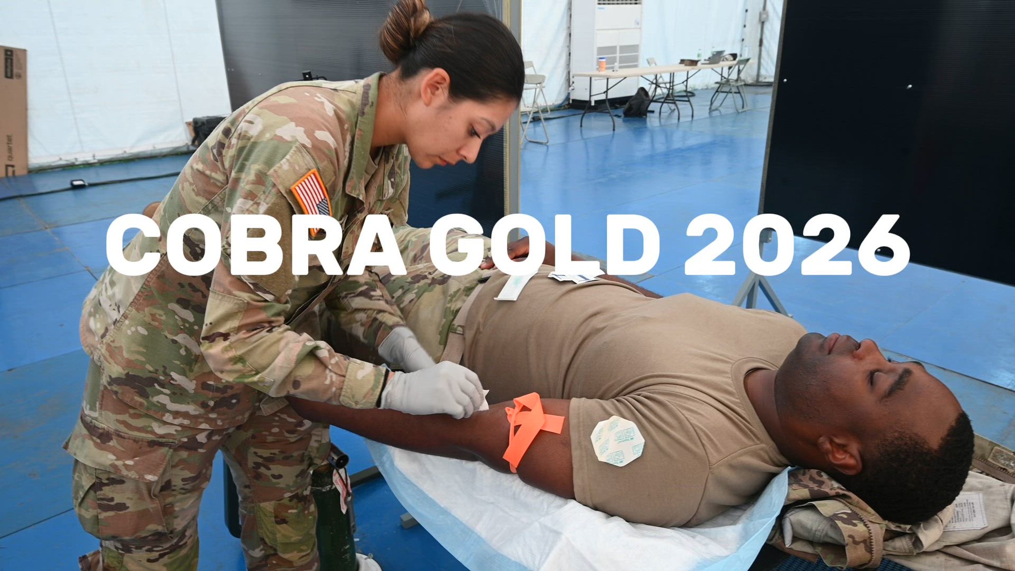 A solider simulates providing medical care to a fellow soldier lying on a cot. 