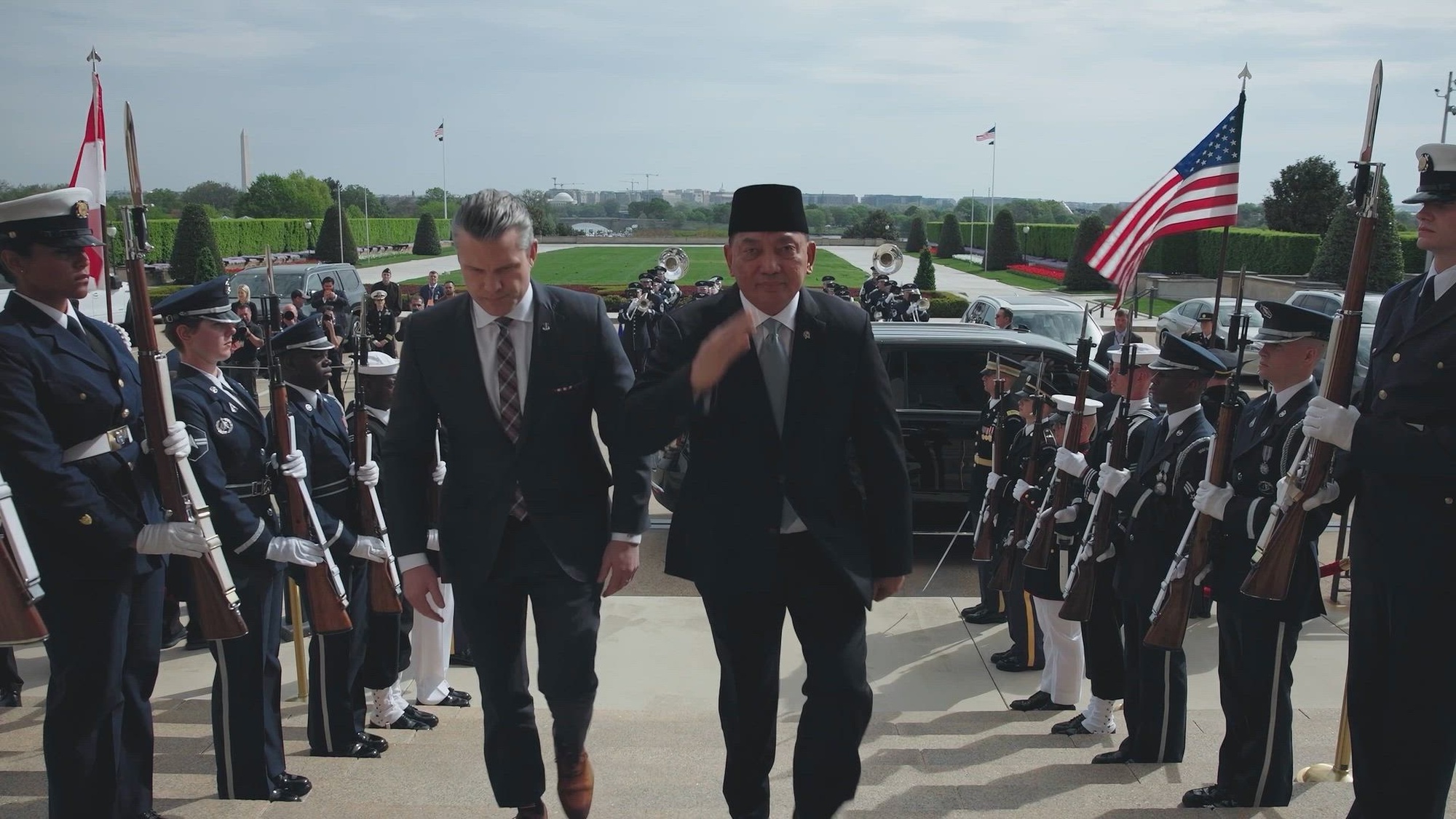 Secretary of War Pete Hegseth and another civilian walk together on a paved surface outdoors, flanked by service members.