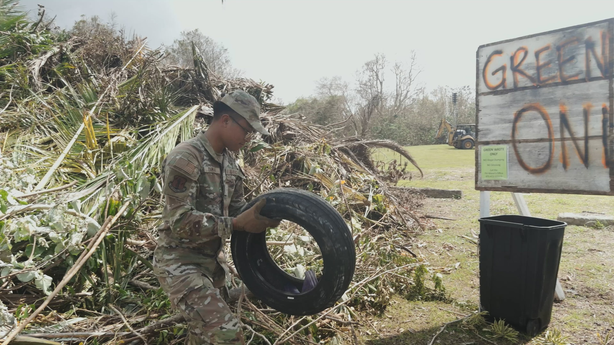 An airman picks up a car tire, with a pile of branches on the ground in the background.