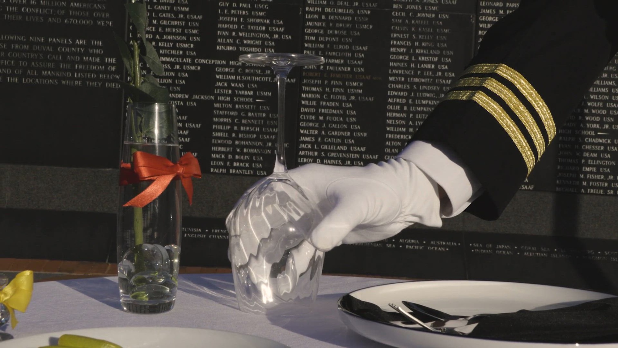 A white-gloved hand places a wine glass on a set table in front of a tablet displaying names.