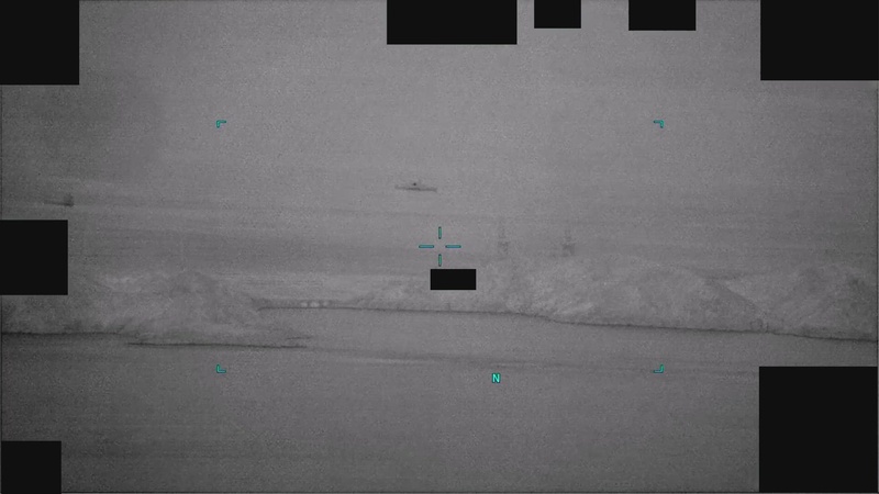 A still frame from the second record, DOW-UAP-PR142, the fourteen second infrared clip. Greyscale thermal imagery of a shoreline with terrain behind it and a centred crosshair reticle. The eight or so solid black rectangles around the edges are official redactions covering the sensor's burned-in data readout, consistent with the release manifest flagging this record as redacted.
