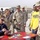A civilian man wearing sunglasses and a dark tracksuit is seated at a decorated table signing a football for military personnel in desert camouflage uniforms. The setting appears to be a military base, likely overseas, with portable structures visible in the background. Several soldiers are gathered around the table, smiling and waiting for autographs.