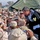 A civilian man in a black jacket with white roses on his shirt leans toward a large crowd of U.S. military personnel in desert camouflage uniforms. The setting appears to be a military base or forward operating location, likely during a USO-style entertainment or morale visit. Soldiers are taking photographs with small digital cameras and appear enthusiastic and engaged.