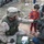 A U.S. Army soldier in desert camouflage uniform is kneeling on the ground providing medical aid to a civilian in a debris-strewn environment. A young child in red pajamas stands nearby observing. The setting appears to be a conflict or disaster zone with rubble, damaged vehicles, and scattered debris visible in the background.