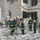 The image shows a large group of emergency responders and firefighters working through the rubble of a severely damaged multi-story building. Personnel wearing yellow and silver helmets with high-visibility uniforms are conducting search and rescue operations amid extensive concrete and brick debris. At least one armed military or security personnel is visible on the right side of the scene.