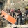 The image shows emergency responders and military personnel working amid rubble and debris, consistent with a post-explosion or structural collapse scene. Firefighters, soldiers, and civilians are collaborating to clear debris and carry what appears to be a stretcher or panel. The scene suggests an active rescue and recovery operation in an urban environment with significant structural damage visible in the background.