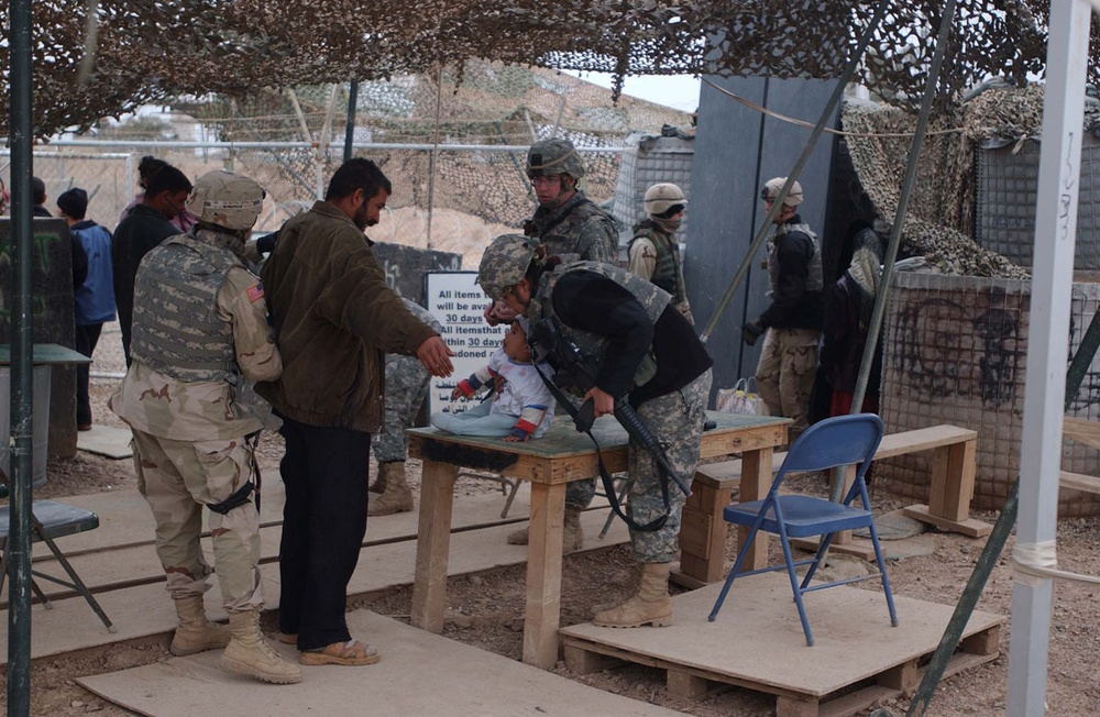 The image shows U.S. military personnel conducting what appears to be a checkpoint or entry control point inspection. Soldiers in ACU (Army Combat Uniform) are examining items on a wooden table while interacting with a civilian. The setting appears to be a forward operating base or security checkpoint in a desert environment, likely in Iraq or Afghanistan.