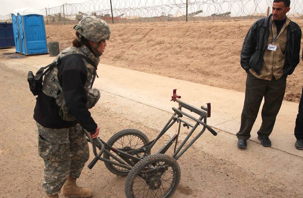 A uniformed soldier wearing ACU (Army Combat Uniform) and helmet is examining a disassembled or stripped bicycle frame/cart on a paved surface at what appears to be a military forward operating base. A civilian male wearing a badge/lanyard stands nearby observing. The background shows concertina wire perimeter fencing, portable toilets, and arid terrain consistent with a Middle Eastern military installation.