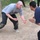Two men are engaged in a hand-to-hand combat training exercise on a dirt surface outdoors. The man on the left is wearing a gray 'Army' PT shirt and black pants with visibly dirty/dusty clothing, suggesting active sparring. Additional observers are visible in the background, indicating this is a supervised training session.