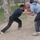 Two men are engaged in a grappling or martial arts sparring session on a dirt surface outdoors. Both are in a clinch position, gripping each other's clothing, with wide stances indicating dynamic movement. Observers are visible in the background near a building and trees.
