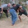 The image shows an outdoor combatives or martial arts training session taking place on a dirt surface near trees and a concrete wall. Two individuals are actively grappling in the center while a third person in a gray shirt observes and appears to be coaching or waiting to participate. Additional observers are visible in the background, including one person in military camouflage uniform.