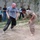 A U.S. Army soldier in a gray 'ARMY' t-shirt engages in a friendly wrestling or grappling match with a local civilian on a dirt surface. Several onlookers, whose faces have been blurred, watch the activity in the background near a concrete wall and eucalyptus tree. The setting and attire suggest an informal cultural exchange or goodwill engagement.