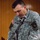 A U.S. Army soldier named Sprecher, wearing Army Combat Uniform (ACU) with the rank of Captain (two silver bars visible), is standing at a microphone podium appearing to speak or read. He is wearing the 101st Airborne Division patch on his left shoulder and a U.S. flag patch on his right shoulder. A cross insignia is visible on his uniform, indicating he may be a military chaplain.
