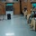 A military briefing or presentation is taking place in what appears to be a dining facility or meeting room on a military installation. A soldier stands at a wooden podium with a microphone, addressing a seated audience of uniformed military personnel. A second person operates a laptop at a side table, likely managing presentation materials for a large display screen in the background.
