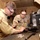 Two U.S. military personnel in desert camouflage uniforms are working together at a workbench inside a plywood-constructed workspace, performing maintenance or repair on a disassembled firearm. Both individuals are wearing protective safety eyewear and appear focused on the task. The setting appears to be a forward operating base or field maintenance facility.
