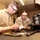 Two U.S. military personnel in desert camouflage uniforms are performing weapons maintenance in what appears to be a field workshop or armory. Both individuals are wearing protective eyewear and are focused on cleaning or inspecting firearm components on a workbench. The setting is a plywood-walled structure consistent with a forward operating base or military installation.