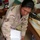 A U.S. Air Force service member wearing desert camouflage (DCU) uniform with the name tape 'LITTLEBULL' is leaning over and pointing to items in a binder. The binder appears to contain handwritten letters and a colorful card reading 'Big Baby Star'. The setting appears to be an indoor common area or recreation room at a military installation.