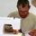 A middle-aged man wearing glasses and a light olive/tan t-shirt is operating a white sewing machine. He is focused intently on guiding fabric through the machine, with a blue object (possibly scissors or a seam ripper) also visible near the work area. The setting appears to be a simple indoor workspace with a white wall in the background.