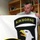 A middle-aged man in a gray t-shirt holds up a large white fabric banner or flag featuring two military unit insignia. The primary insignia is the 101st Airborne Division patch (Screaming Eagle), and a second shield-shaped patch is partially visible in the lower left. The setting appears to be an indoor room with bookshelves in the background.