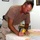 A middle-aged man wearing a tan t-shirt and military-style camouflage pants is leaning over a table engaged in fabric cutting or quilting. He is using a yellow rotary cutter and a clear quilting ruler on a cutting mat, with various pieces of fabric visible on the table. The setting appears to be an indoor recreational or common room, with framed artwork on the walls and a chess set visible in the background.