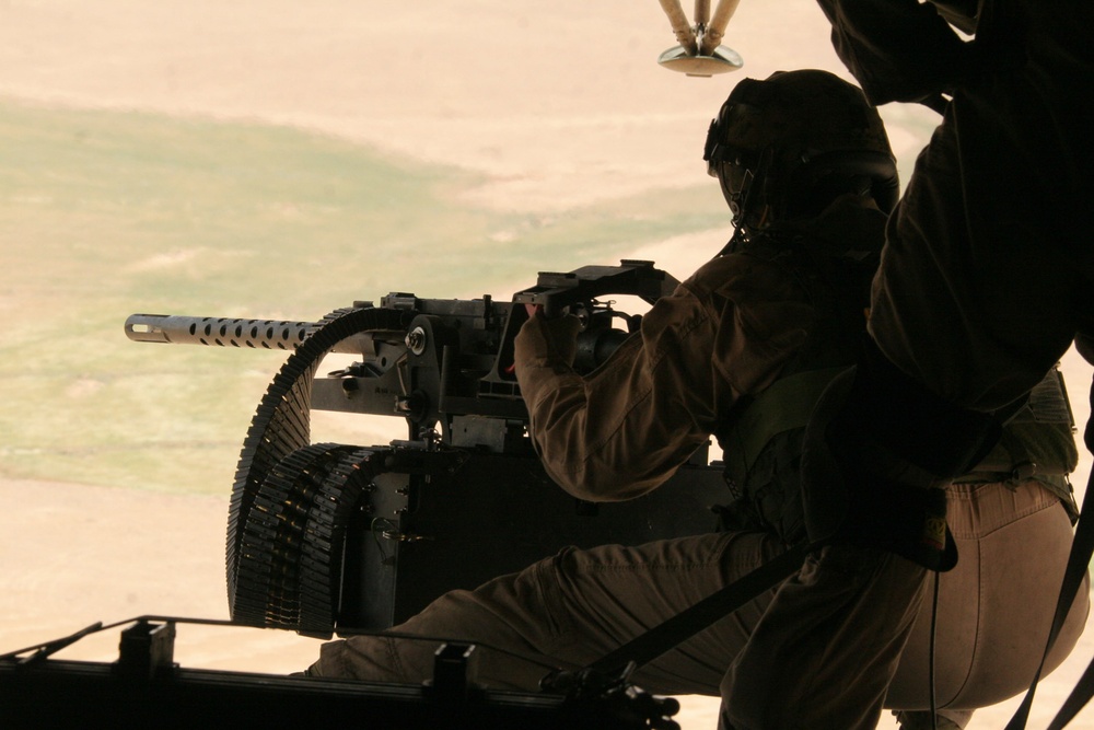 A military door gunner is positioned at a helicopter-mounted machine gun, viewed from inside the aircraft. The gunner is wearing a flight helmet and military uniform, operating what appears to be a belt-fed machine gun with a visible ammunition belt. The ground terrain below is arid and sandy, consistent with a desert environment.