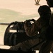 A military door gunner is positioned at a helicopter-mounted machine gun, viewed from inside the aircraft. The gunner is wearing a flight helmet and military uniform, operating what appears to be a belt-fed machine gun with a visible ammunition belt. The ground terrain below is arid and sandy, consistent with a desert environment.
