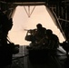 The image shows military personnel operating a crew-served weapon system from the open rear ramp of a large military helicopter, likely a CH-47 Chinook. The figures are silhouetted against a bright, hazy exterior sky, creating a high-contrast composition. At least two personnel are visible manning what appears to be a mounted machine gun at the ramp opening.