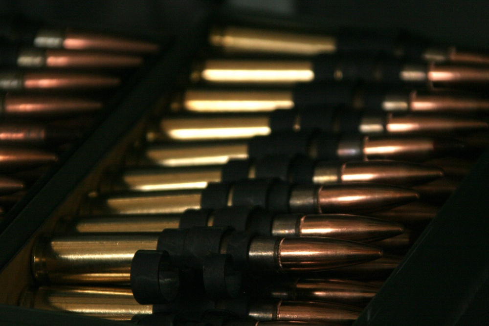 The image shows a close-up view of linked ammunition rounds arranged in belt-feed configuration, typical of machine gun ammunition. The cartridges display copper-jacketed projectiles and brass casings connected by dark metal links. The photograph is taken at a shallow depth of field, with foreground rounds in sharper focus and background rounds progressively blurred.