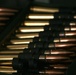 The image shows a close-up view of linked ammunition rounds arranged in belt-feed configuration, typical of machine gun ammunition. The cartridges display copper-jacketed projectiles and brass casings connected by dark metal links. The photograph is taken at a shallow depth of field, with foreground rounds in sharper focus and background rounds progressively blurred.