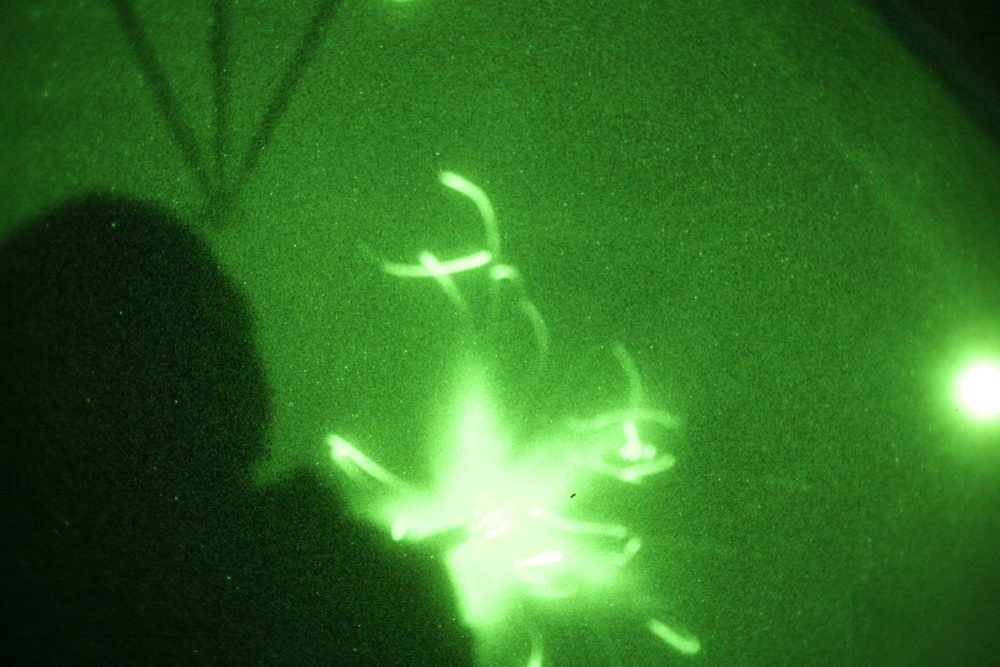 This image was captured through night vision optics, rendering everything in characteristic green monochrome. A small multi-rotor drone (quadcopter) is visible in flight, with its propellers appearing as bright, motion-blurred arcs due to their rotation speed. A human silhouette is partially visible on the left side of the frame, likely the operator or an observer.