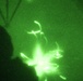 This image was captured through night vision optics, rendering everything in characteristic green monochrome. A small multi-rotor drone (quadcopter) is visible in flight, with its propellers appearing as bright, motion-blurred arcs due to their rotation speed. A human silhouette is partially visible on the left side of the frame, likely the operator or an observer.