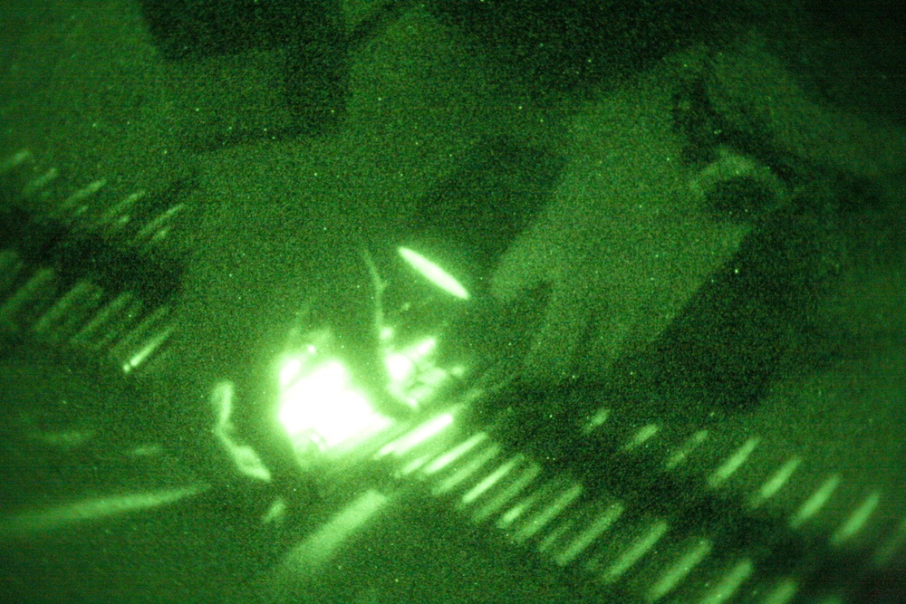 This image was captured through night vision optics, producing the characteristic green monochromatic appearance. A bright central light source dominates the frame, surrounded by blurred and grainy elements suggesting motion or low-light conditions. The image quality is heavily degraded due to the night vision amplification and apparent camera movement.