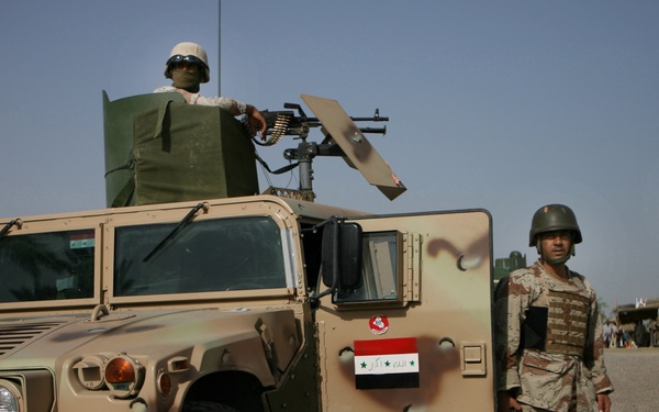 The image shows military personnel alongside a tan/desert-colored HMMWV (Humvee) in what appears to be a Middle Eastern operational environment. An Iraqi flag decal is visible on the vehicle door, and soldiers in different uniforms are interacting near the vehicle. One soldier is positioned in the turret manning a crew-served weapon.