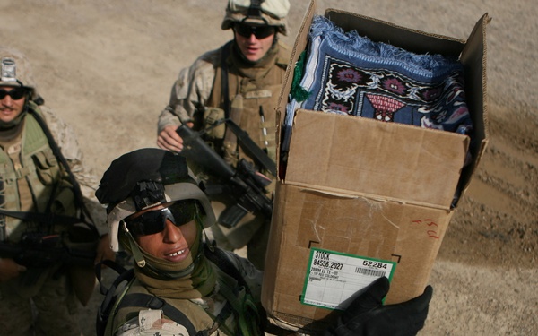 Three military personnel in desert camouflage uniforms are engaged in what appears to be a supply or humanitarian aid distribution operation. One soldier in the foreground is carrying a large cardboard box containing folded textiles or blankets toward a military vehicle. Two armed soldiers stand in the background providing security or oversight.