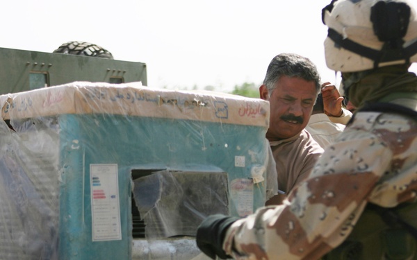 The image shows military personnel and a civilian collaboratively carrying a large appliance, likely an air conditioning unit or generator, still wrapped in plastic packaging. Two soldiers in desert camouflage uniforms are working alongside a civilian man in traditional Middle Eastern attire. The setting appears to be an outdoor area with military vehicles visible in the background.