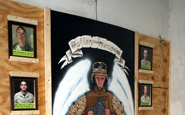 A military memorial display titled 'Fallen Heroes' consisting of a large painted mural mounted on plywood, flanked by framed portrait photographs of fallen service members. The central painting depicts a soldier in desert camouflage with angel wings, holding a shield inscribed with names and dates, with a banner reading 'Freedom Shield' and text 'Ready to Fight'. The display appears to be located inside a military installation, likely in a combat zone given the construction materials and context.